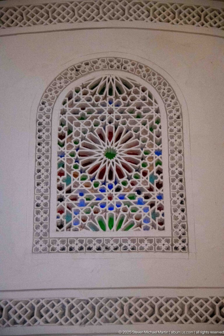 Arched white plaster grille with radial geometric pattern and small blue, green, red, and amber glass accents behind.