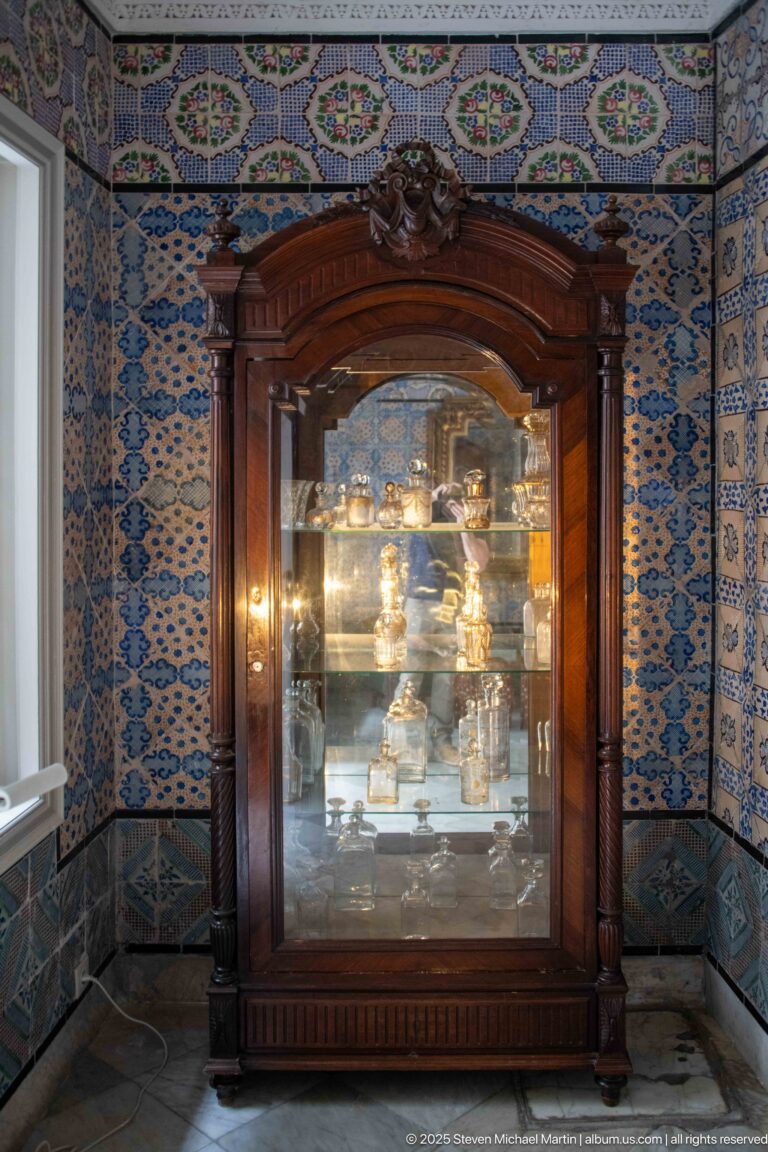 Dark carved wooden glass-front cabinet displaying metal objects against blue-and-white tiled walls.