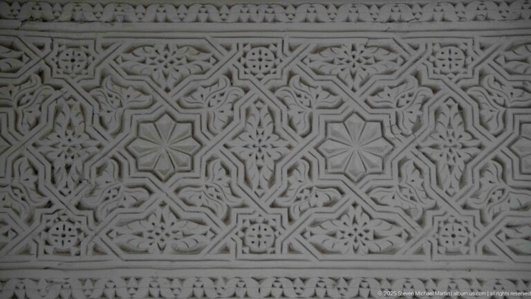 Horizontal pale plaster frieze with dense interlacing geometry, repeated rosettes, stars, and narrow carved borders.