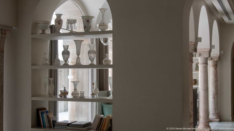 White arched shelving with ceramics, glass vessels, and books, opening toward pale stone columns in the hall beyond.