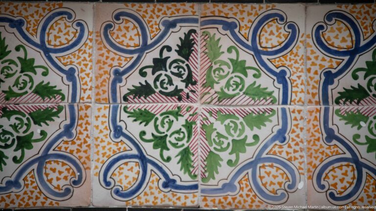Close view of four ceramic tiles with blue scrolls, green foliage, pink hatching, and ochre accents forming a symmetrical pattern.