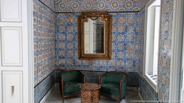 Small tiled sitting room with gilded mirror, two green chairs, inlaid table, and daylight from a side window.