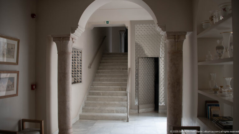 Marble staircase in an arched hall with pale stone columns, pierced screen, framed art, and built-in display shelves.