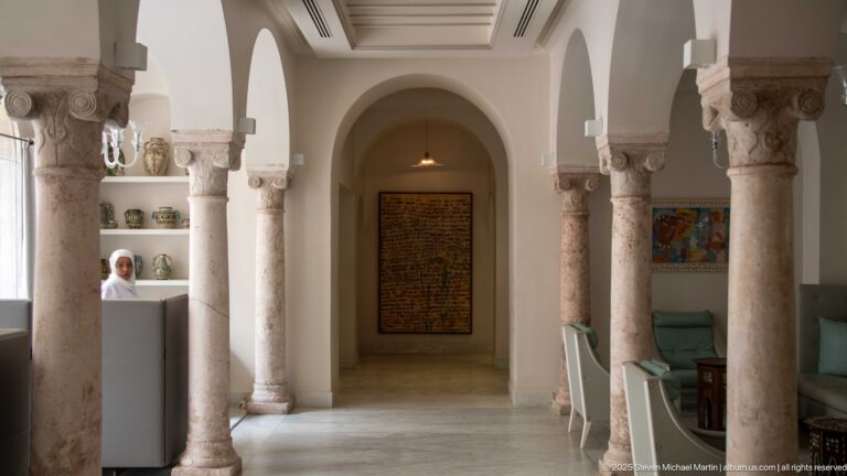 Arcaded interior corridor with pale stone columns, display shelves, seating, and framed artwork at the far end.