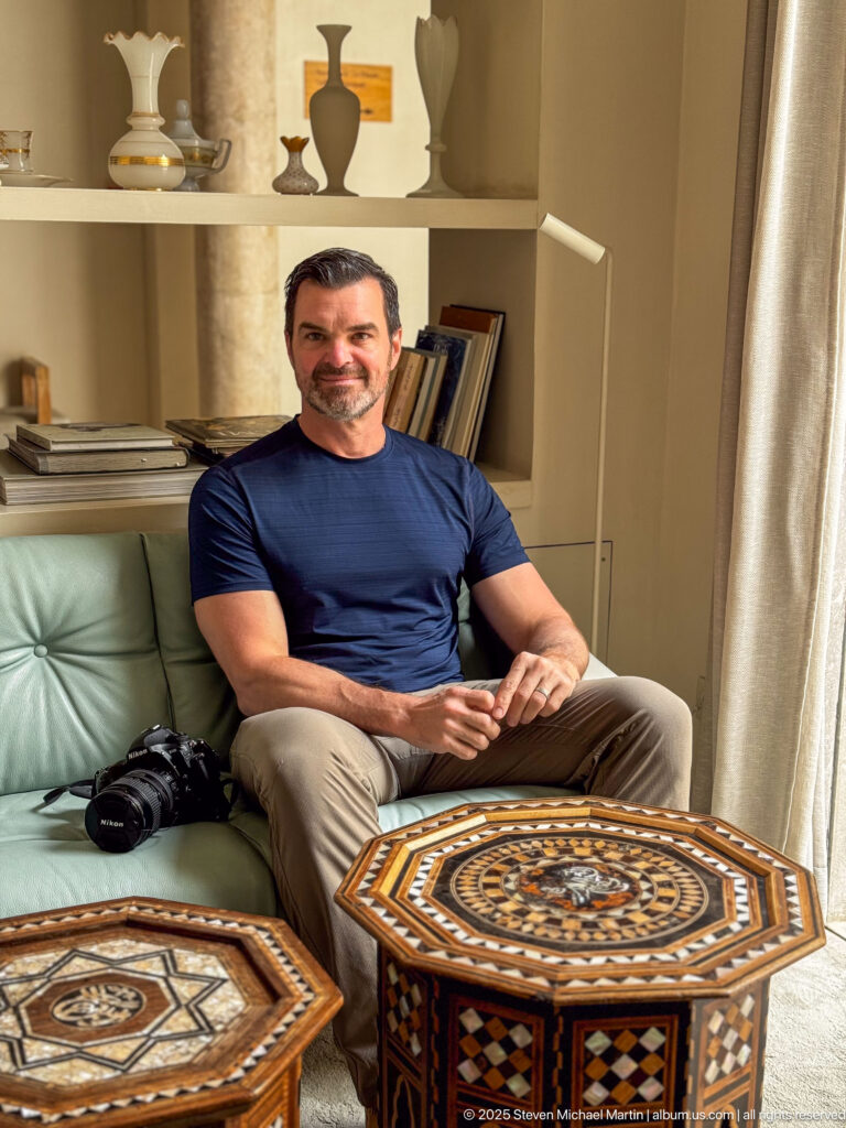 Seated man on pale green sofa beside two octagonal inlaid wooden tables, with books and display objects behind.