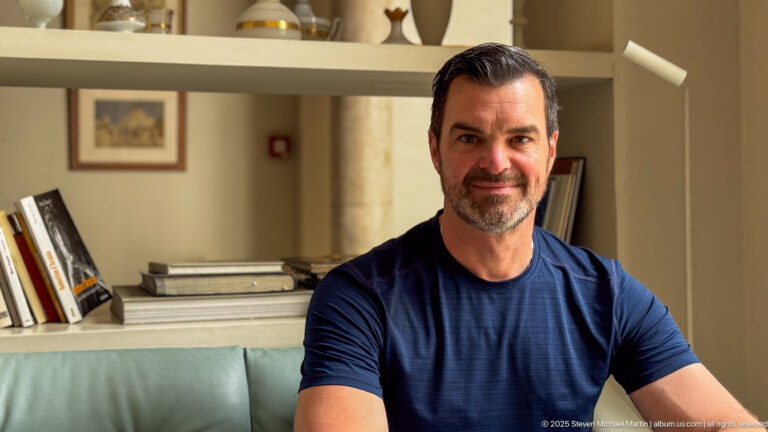 Close portrait of a seated man in a navy shirt, with pale green sofa, books, and decorative shelving behind.