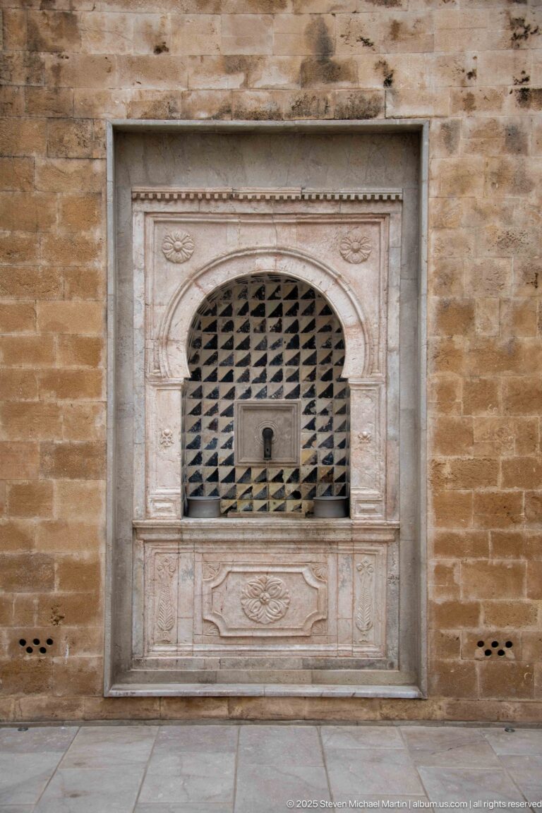Arched dark lattice opening within a carved pale stone frame set into a warm masonry wall.