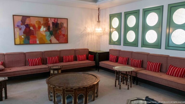 Long lounge with brown upholstered benches, red striped cushions, inlaid tables, green circular-screen shutters, and modern artwork.