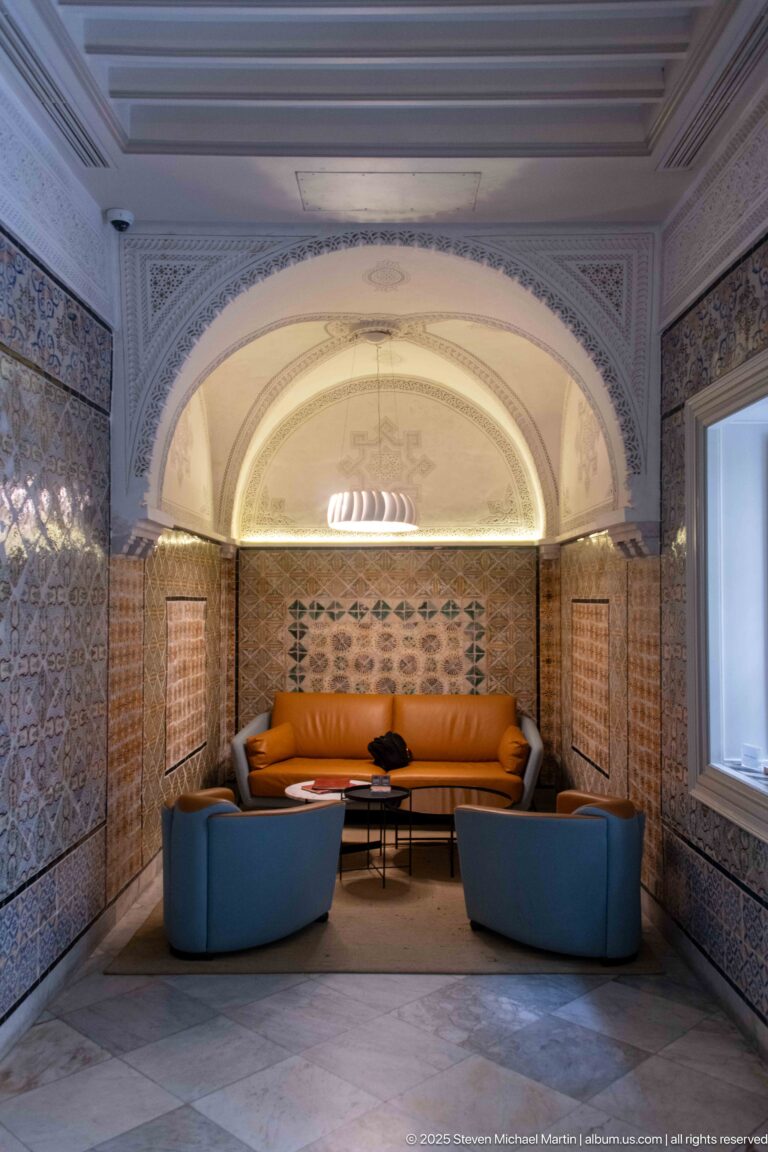 Symmetrical lounge with orange sofa and blue chairs set beneath a broad arch and surrounded by patterned tilework.