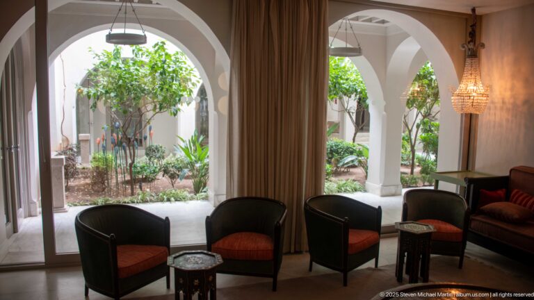 Dark lounge chairs and small inlaid tables framed by broad arches opening onto a bright planted courtyard.