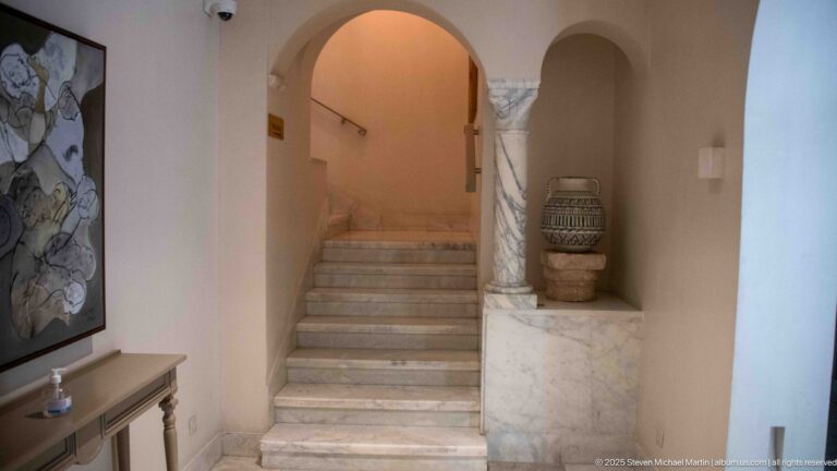 Marble stairs rise beneath rounded arches beside veined stone facing, a carved column, wall art, and a large ceramic vessel.