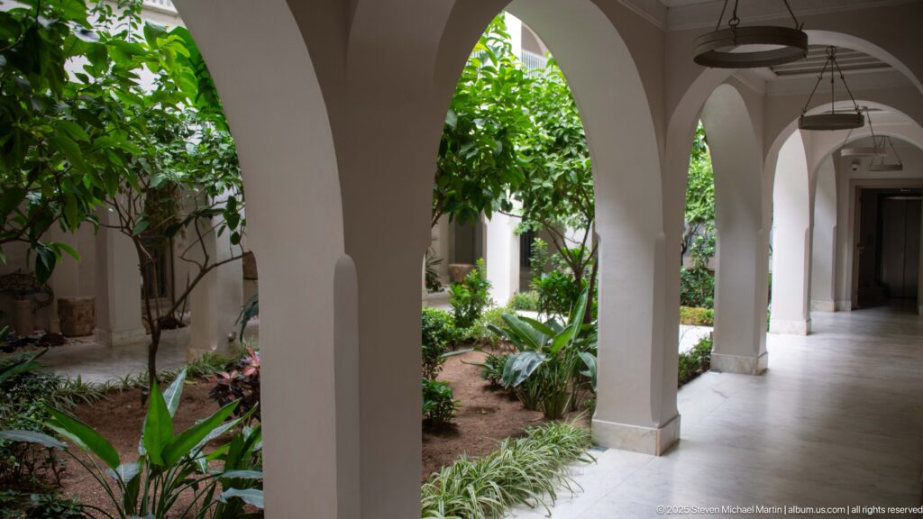 White arcades open onto a lush interior garden with trees and tropical foliage beside a tiled covered walkway.