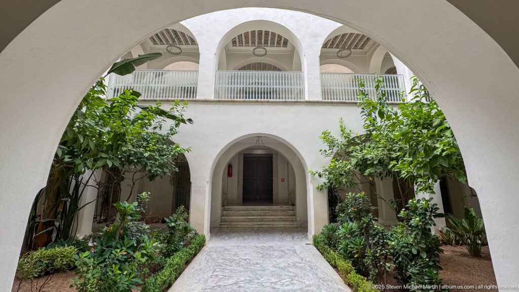 View through a large arch into a planted courtyard with a central path and three-arched upper gallery.