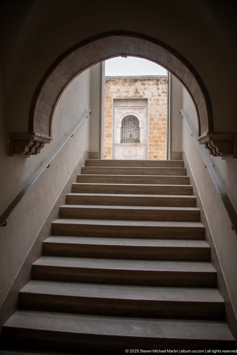 Wide staircase rises through a massive stone arch toward a lit landing with a recessed decorative panel.