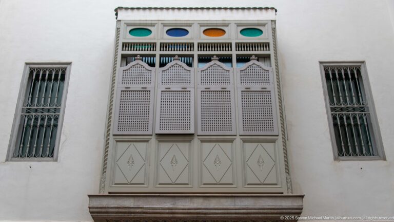 Tall pale wooden screened window with geometric panels and colored glass, flanked by two narrow barred windows.