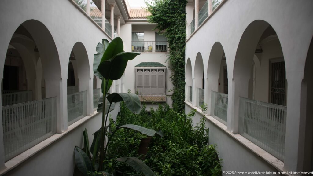 Two stories of white arcades surround a narrow planted courtyard with tall foliage, vines, and a screened window.