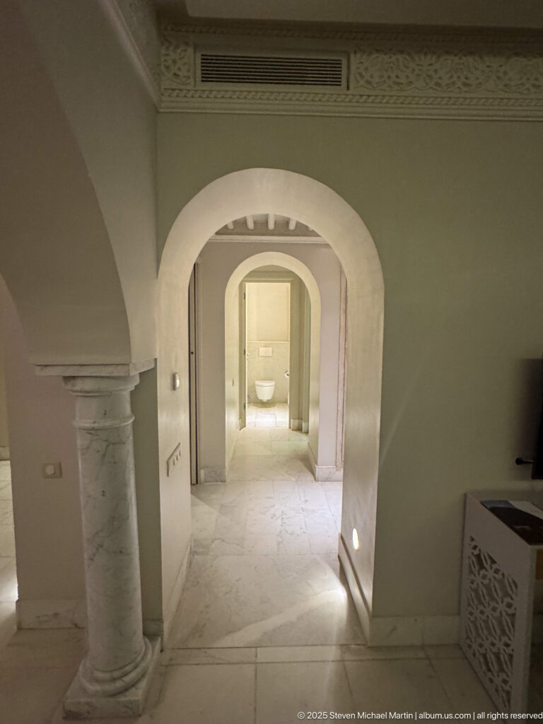 Pale interior passage with nested arches, short stone columns, marble floor, and a brightly lit room beyond.