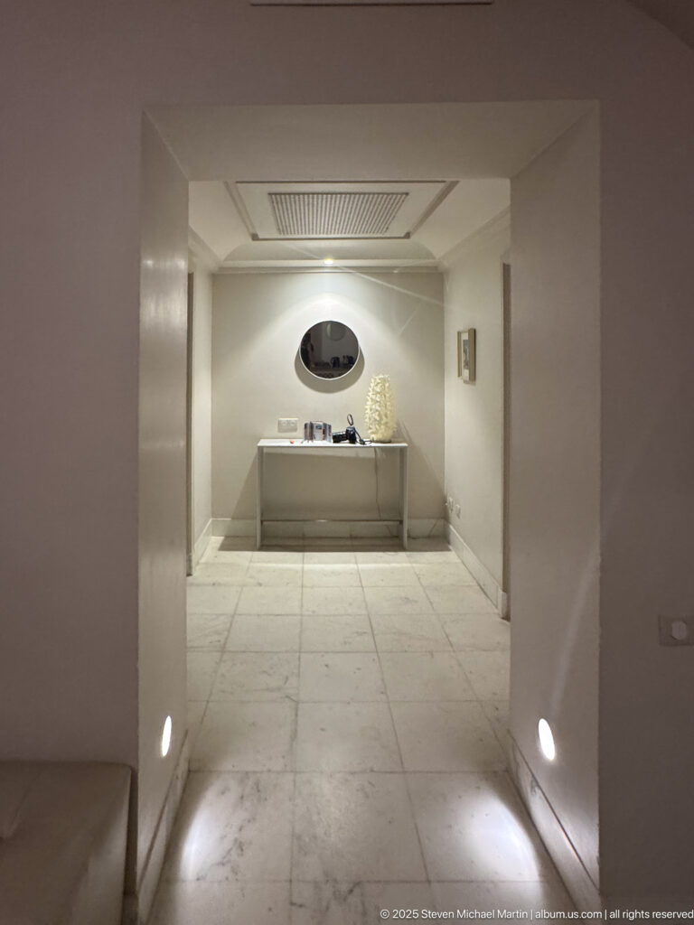 Minimalist pale vestibule with marble floor, low wall lights, an oval mirror, console, and ceiling grille.