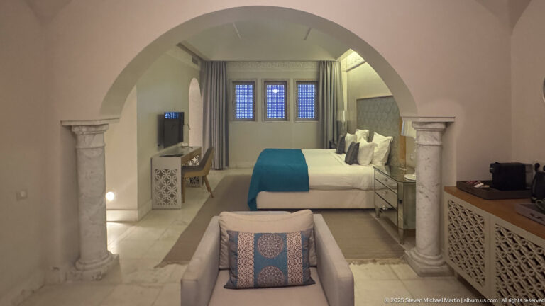 Guest bedroom seen through a large arch with stone columns, blue bed runner, armchair, and lattice-front cabinetry.