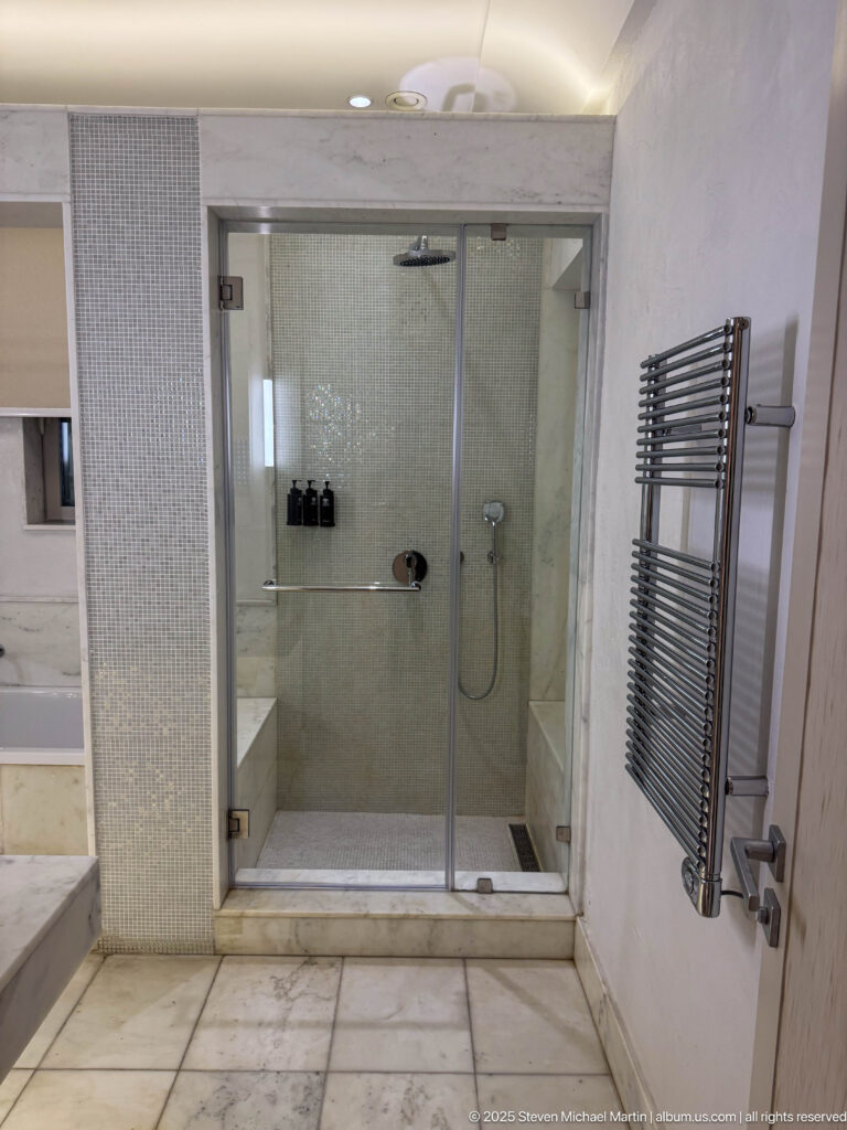Glass walk-in shower with pale mosaic walls, bench, shower fittings, marble floor, and chrome towel warmer.
