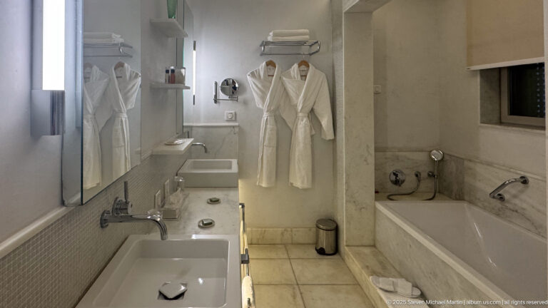 Long marble bathroom with two rectangular sinks, a soaking tub, white bathrobes, towels, and wall mirror.