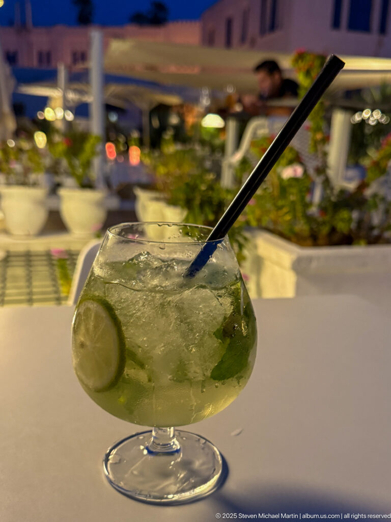Stemmed glass of pale green drink with lime slices, mint leaves, ice, and black straw on an evening terrace.