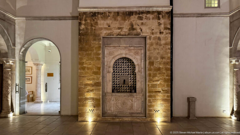 Illuminated carved wall niche with perforated arch set into rough stone masonry beside a rounded passage.