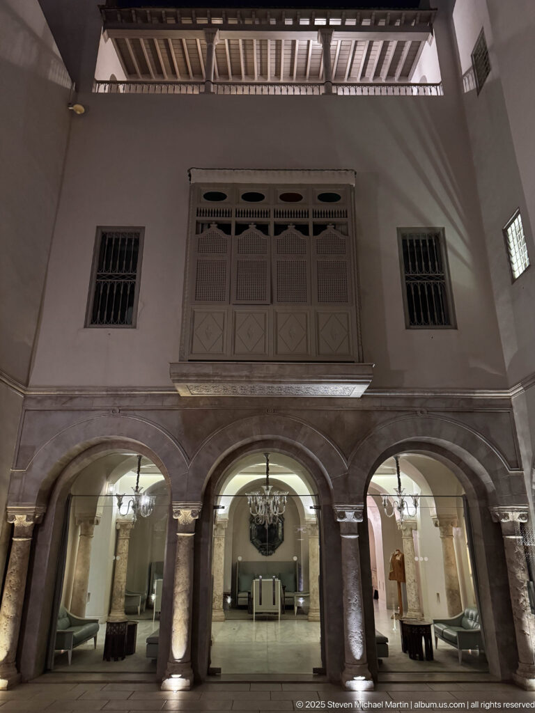 Night view of three illuminated courtyard arches beneath a projecting screened window and exposed roof beams.