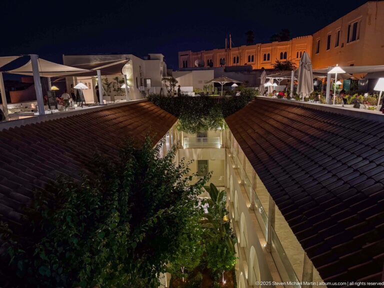 Evening rooftop terrace with tables and umbrellas overlooking tiled roofs, greenery, and the illuminated inner courtyard.