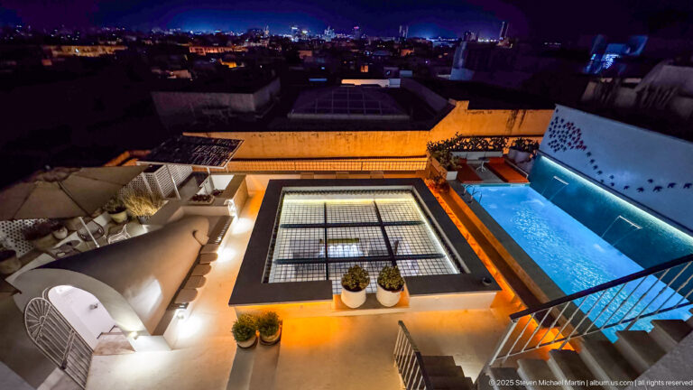 Nighttime rooftop terrace with a bright blue plunge pool, lounge seating, central gridded skylight, and city roofs beyond.