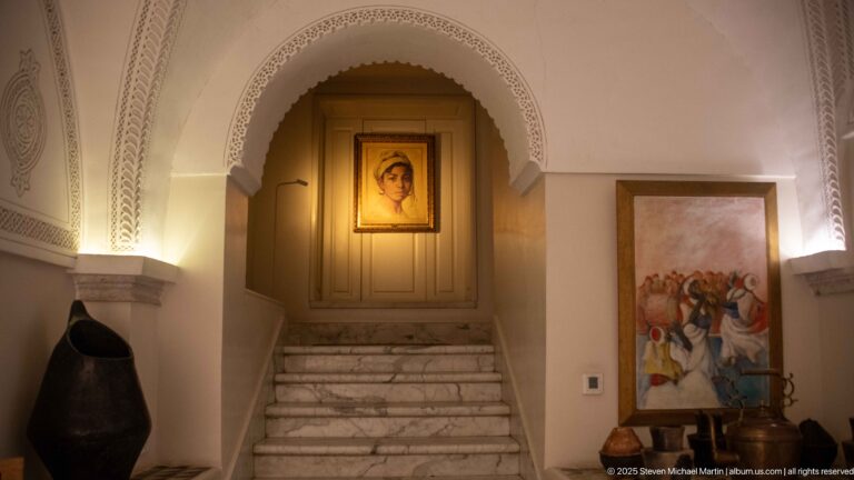Marble stair beneath an ornate pointed arch, with pale carved plaster, framed artwork, metal vessels, and warm lighting.