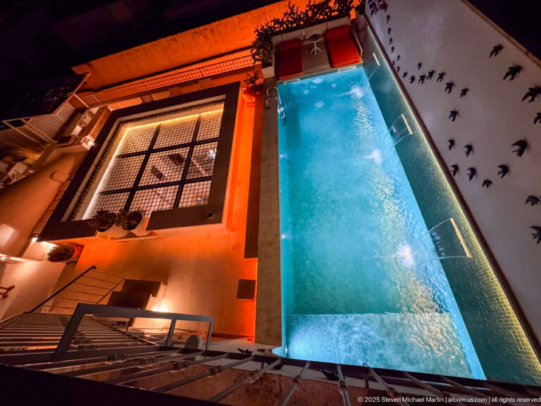 Overhead night view of a narrow blue rooftop pool beside amber-lit walls, railing, planters, and a gridded skylight.