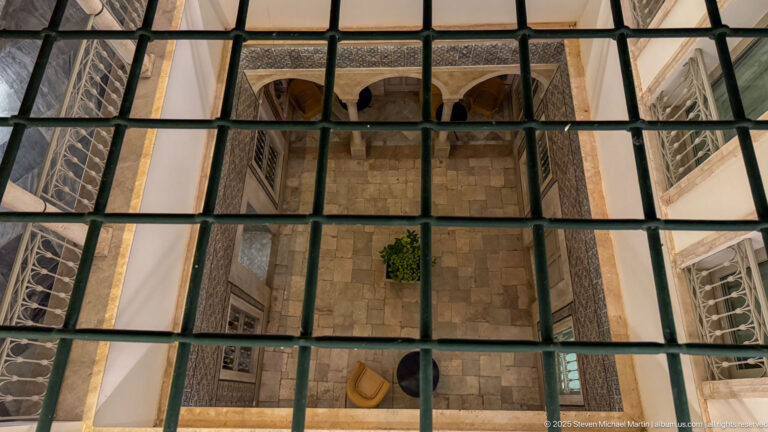 Grid of glass panes viewed from above with a stone-paved courtyard, arches, windows, and seating visible below.