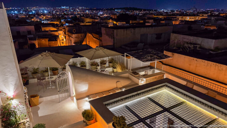 Night rooftop terrace with glowing gridded skylight, outdoor seating, plants, and dense city rooftops beyond.
