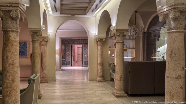 Interior gallery with carved stone columns, rounded arches, pale floor, lounge furnishings, and patterned doorway beyond.