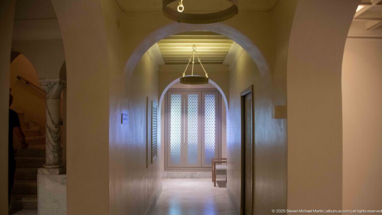 Long pale corridor with rounded arches, hanging circular lights, side doorways, and screened double doors at the far end.