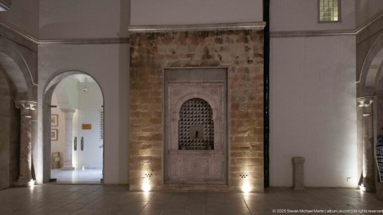 Carved rectangular niche with small perforated arch centered in an uplighted exposed stone wall between two passages.