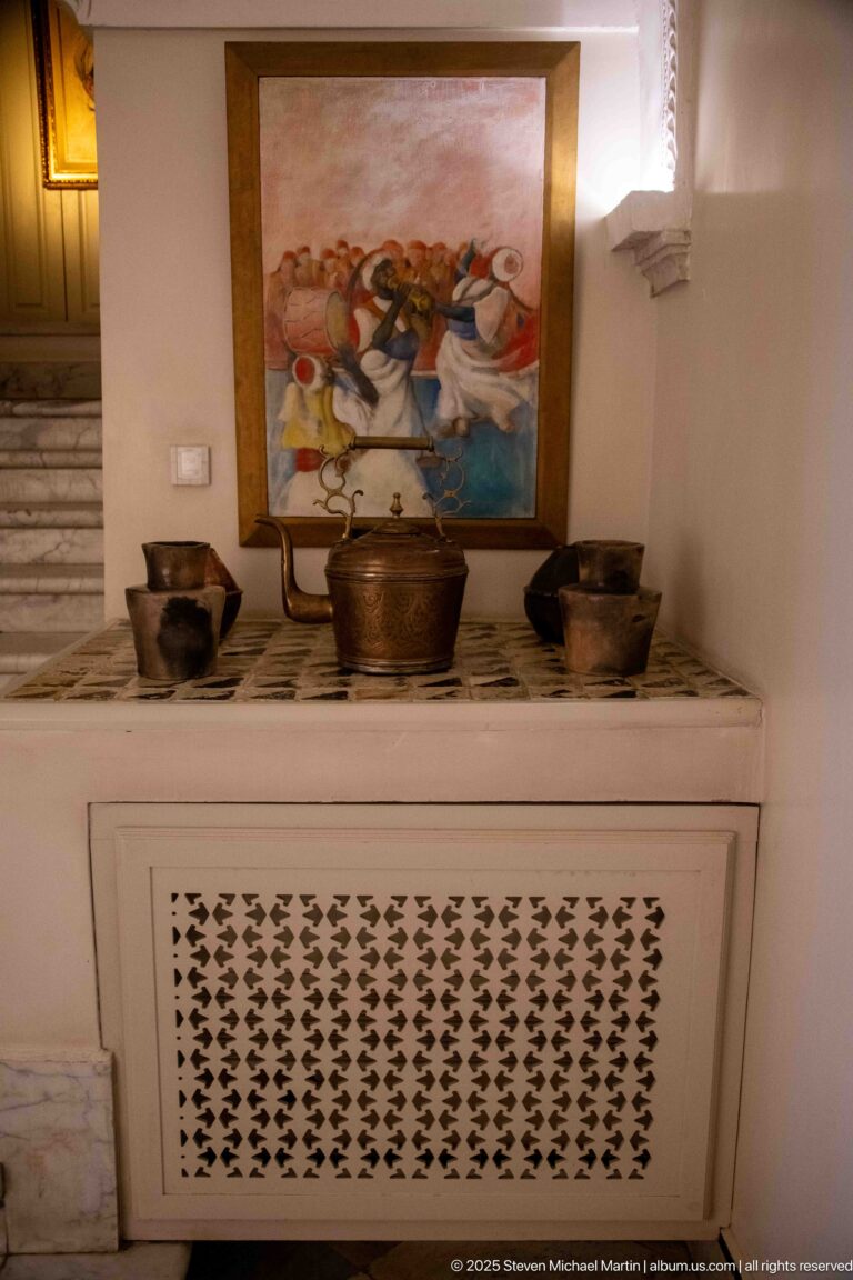 Metal bowls and vessels arranged on a marble ledge beneath a colorful painting and above a perforated white grille.