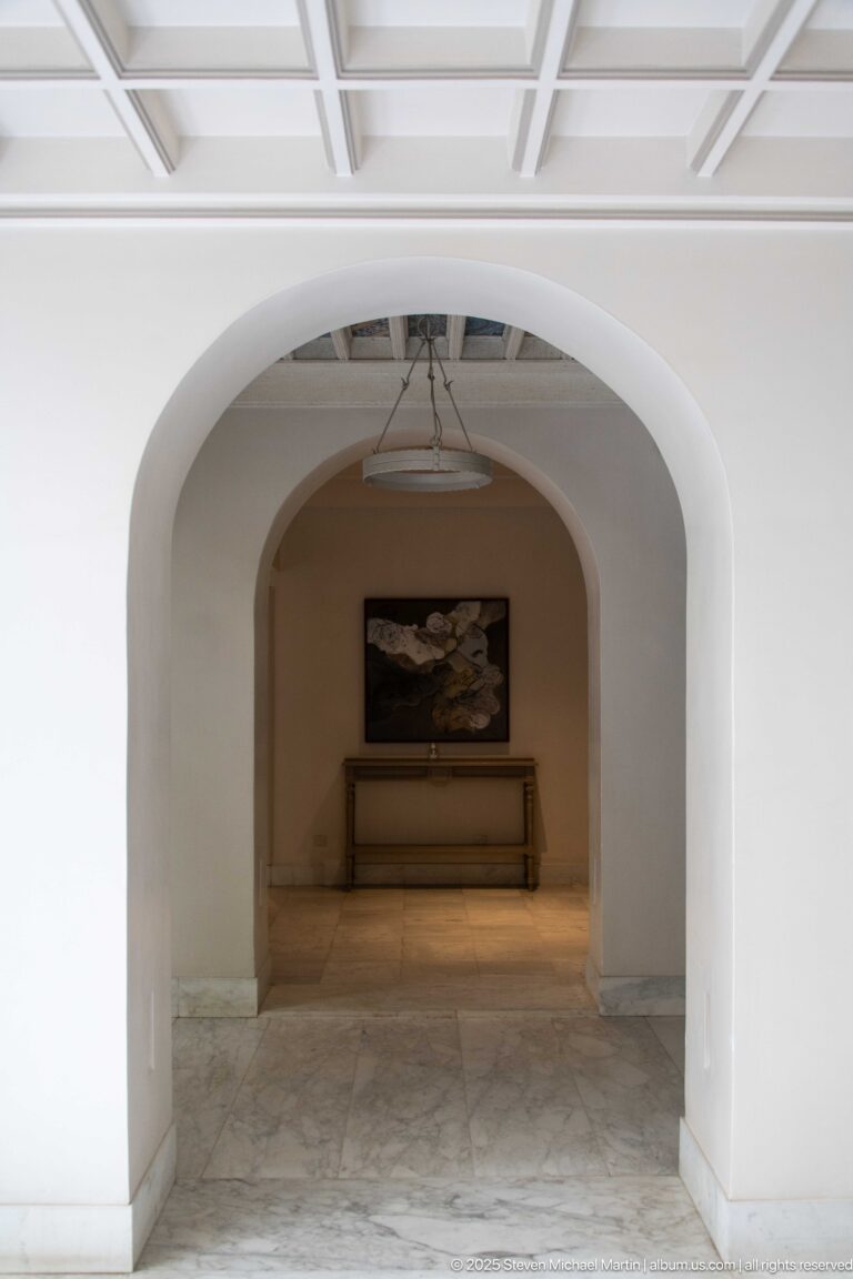 Large white arch opens into a softly lit room with pale stone floor, console table, framed artwork, and coffered ceiling.