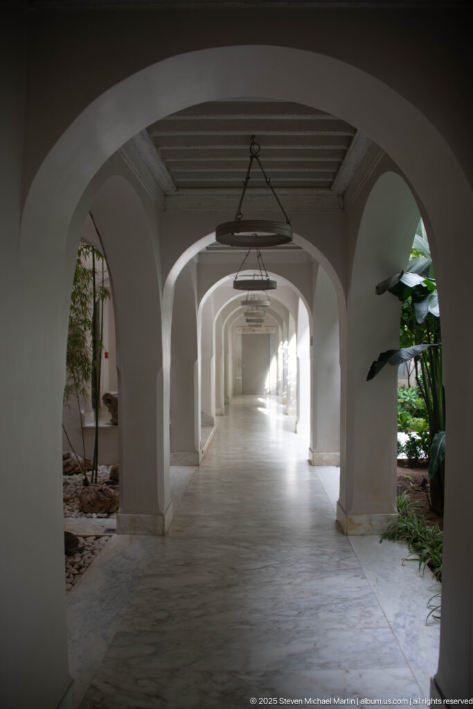 Long white arcade with repeated rounded arches, pale stone floor, hanging circular lights, and planted courtyard openings along both sides.