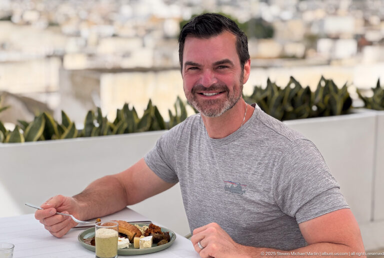 Smiling man seated at an outdoor table with breakfast, backed by white terrace walls, succulents, and blurred city rooftops.