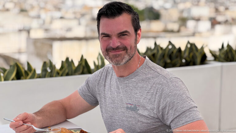 Man in a gray shirt seated at an outdoor breakfast table, with terrace plants and blurred rooftops behind him.