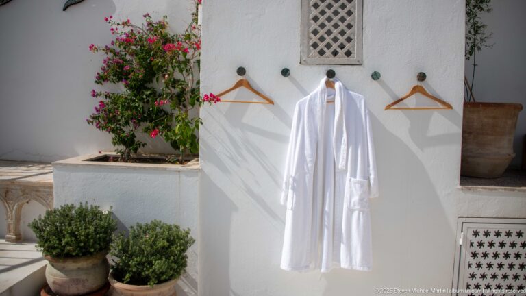 White bathrobe and wooden hangers on a sunlit wall beside potted plants, a small lattice opening, and patterned vent panel.