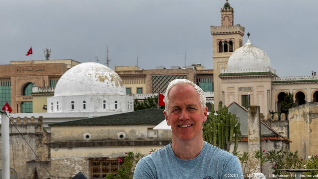 Smiling man on a rooftop terrace with white domes, a square minaret, Tunisian flags, cactus, and historic buildings behind.