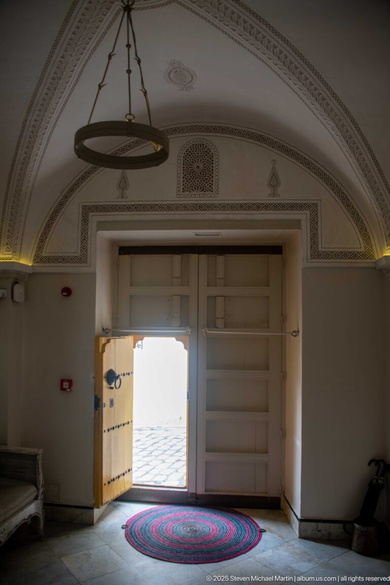 Vaulted pale entrance hall with relief ornament, circular rug, and open yellow studded door leading to a bright courtyard.
