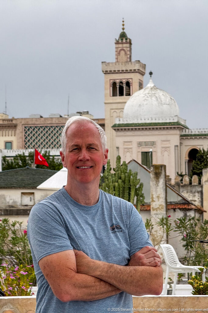Smiling man with folded arms on a rooftop terrace, with a white dome, square tower, Tunisian flag, and plants behind.