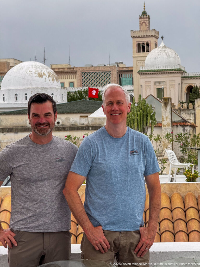Two smiling men standing on a rooftop terrace with white domes, square tower, Tunisian flag, and tiled roofs behind.
