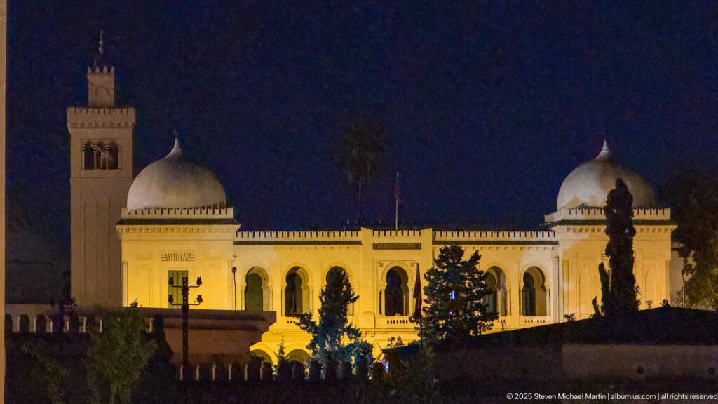 Illuminated pale building at night with two white domes, a square tower, arched windows, trees, and a low balustrade.