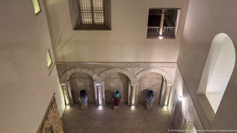 View from above into a stone courtyard with three arches, large ceramic vessels, floor lights, and high surrounding walls.