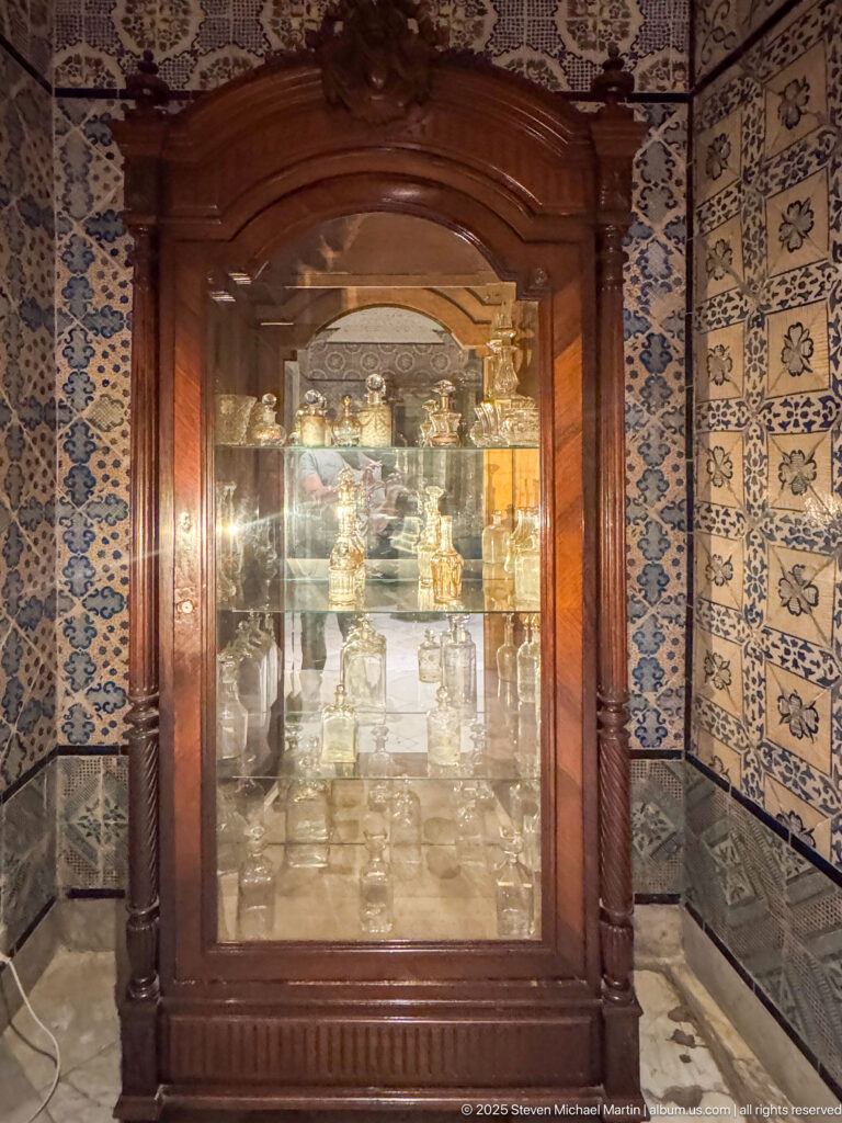 Dark carved wooden display cabinet with glass doors and metal objects, surrounded by blue-and-white patterned tile walls.